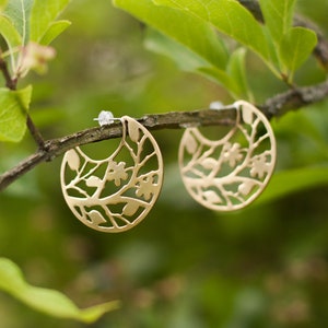 Round brass earrings with tree branches motif cut out are balancing on the real tree branch. Earrings are shiny and golden, but not polished, they have a soft matte finish. Their pattern is tree branches with leaves and flowers.