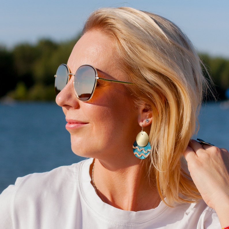 Woman wears earrings, consisting of two circles, one overlaying another. The upper circle is made of brass, the lower one is made of blue patinated copper with 2 polished copper wavy lines. Earrings are long, they hang along the woman's neck.