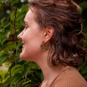 Woman with curly hair is wearing disk earrings of golden metal with natural pattern cut out. The pattern is showing tree branch with leaves and flowers, it reminds of blooming sakura drawing. Earrings are flat with a stud ear wire.