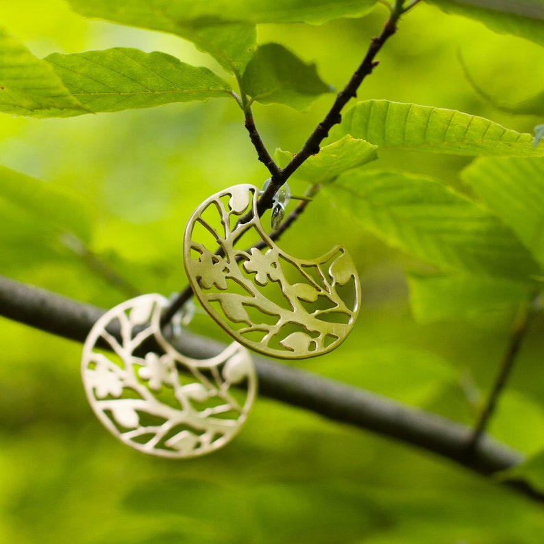 Round brass earrings with tree branches motif cut out are balancing on the real tree branch. Earrings are shiny and golden, but not polished, they have a soft matte finish. Their pattern is tree branches with leaves and flowers.