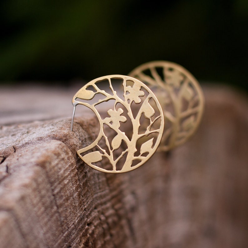 Round brass earrings with tree branches motif cut out are balancing on the round piece of wood. Earrings are shiny and golden, but not polished, they have a soft matte finish. Their pattern is tree branches with leaves and flowers.