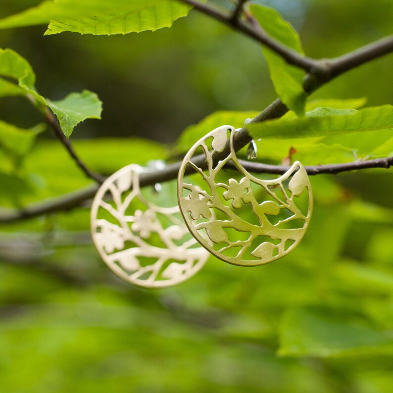 Round brass earrings with tree branches motif cut out are balancing on the real tree branch. Earrings are shiny and golden, but not polished, they have a soft matte finish. Their pattern is tree branches with leaves and flowers.