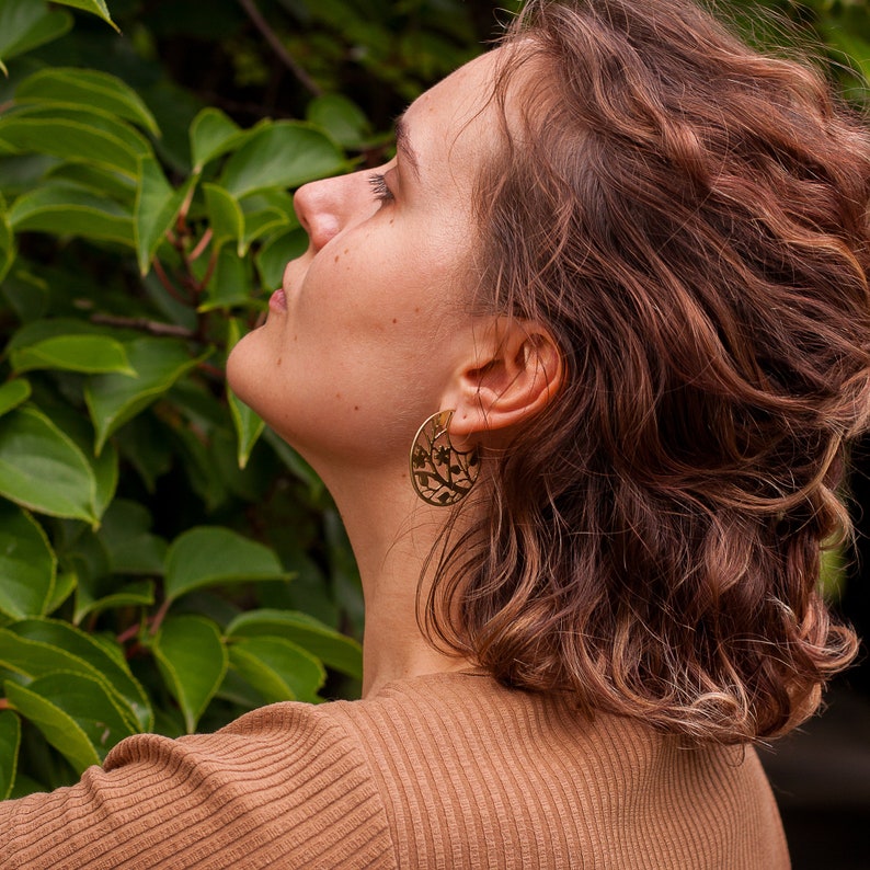 Woman with curly hair is wearing disk earrings of golden metal with natural pattern cut out. The pattern is showing tree branch with leaves and flowers, it reminds of blooming sakura drawing. Earrings are flat with a stud ear wire.