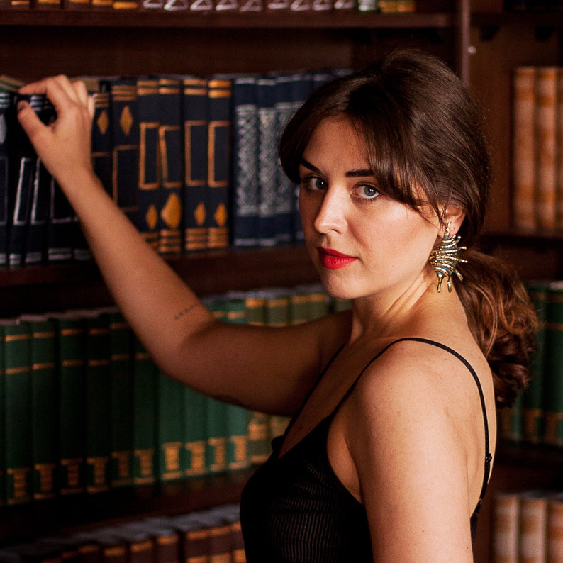 Young woman wearing big golden brass earrings in the form of elongated spikey shells. Earrings are bold and eye-catching, they are long and ribbed.