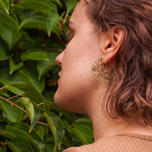 Woman with short hair is wearing disk earrings of golden metal with natural pattern but out. We see earrings from aside, they are flat and all of their pattern is nicely seen. Earrings are flat with a stud ear wire.