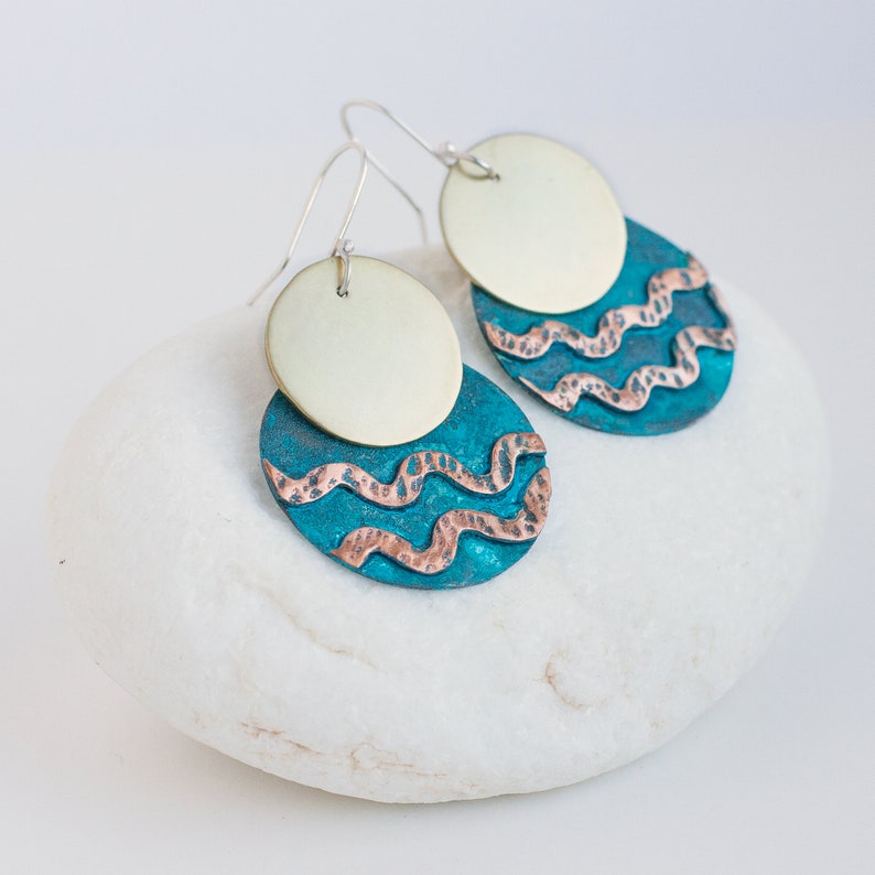 Mixed metal earrings, consisting of two circles, one overlaying the other, lie on the white stone. The upper circle is made of polished brass, the lower one is made of blue patinated copper with 2 hammered copper wavy lines.
