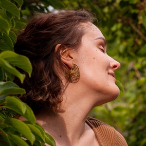 Woman with curly hair is wearing disk earrings of golden metal with natural pattern cut out. The pattern is showing tree branch with leaves and flowers, it reminds of blooming sakura drawing. Earrings are flat with a stud ear wire.