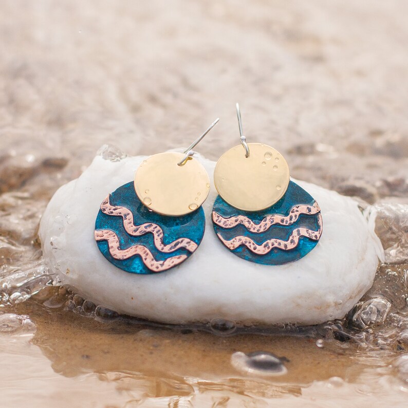 Mixed metal earrings, consisting of two circles, one overlaying the other, lie on the white stone surrounded by water. The upper circle is made of polished brass, the lower one is made of blue patinated copper with 2 polished copper wavy lines.