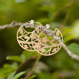 Round brass earrings with tree branches motif cut out are balancing on the real tree branch. Earrings are shiny and golden, but not polished, they have a soft matte finish. Their pattern is tree branches with leaves and flowers.