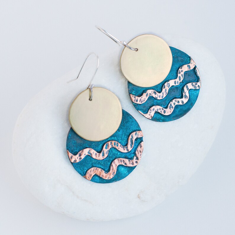 Mixed metal earrings, consisting of two circles, one overlaying the other, lie on the white stone. The upper circle is made of polished brass, the lower one is made of blue patinated copper with 2 hammered copper wavy lines.