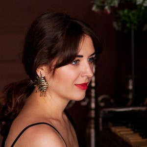 Young woman in bold golden metal earrings in the form of elongated spikey shells. Earrings are made of golden brass with naturally added blue patina.