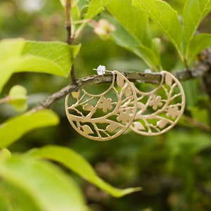 Round brass earrings with tree branches motif cut out are balancing on the real tree branch. Earrings are shiny and golden, but not polished, they have a soft matte finish. Their pattern is tree branches with leaves and flowers.