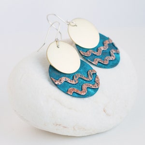 Mixed metal earrings, consisting of two circles, one overlaying the other, lie on the white stone. The upper circle is made of polished brass, the lower one is made of blue patinated copper with 2 hammered copper wavy lines.