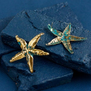Hand-made nrass starfish earrings made os brass with blue patina and silver ear wire. One earring is laying showing the back side. It is seen that the earrings are not flat, thay mimic the real starfish surface.