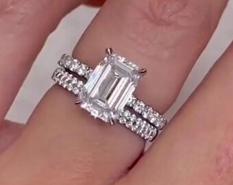 May include: A silver engagement ring set featuring a large, emerald-cut diamond center stone. The ring is accompanied by a matching band, both adorned with smaller, sparkling accent stones. The jewelry is displayed on a person's finger.