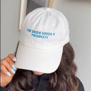 May include: White baseball cap with blue embroidery that reads "The Bride Needs A Prosecco".