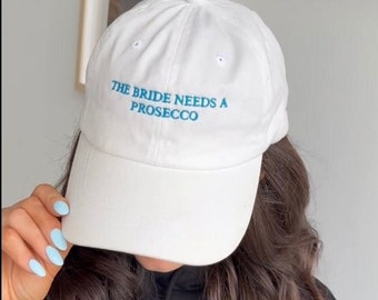 May include: White baseball cap with blue embroidery that reads "The Bride Needs A Prosecco".