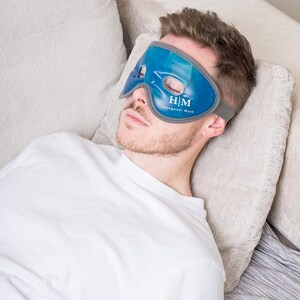 K&ouml;nnte beinhalten: Eine Person tr&auml;gt eine blaue Gel-Augenmaske mit dem Text "H/M Hangover Mask". Die Maske ist dazu gedacht, Kopfschmerzen und Augenbelastung zu lindern.