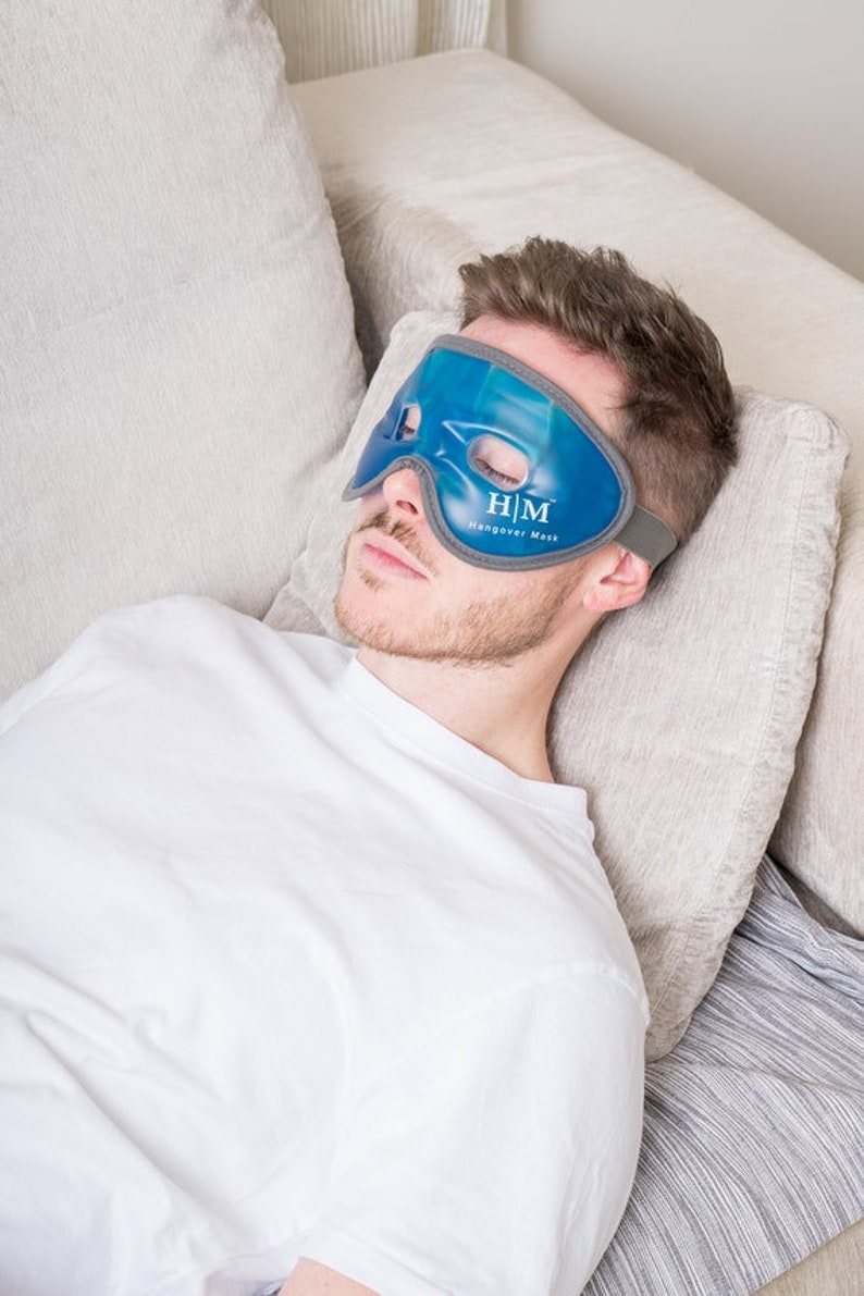 K&ouml;nnte beinhalten: Eine Person tr&auml;gt eine blaue Gel-Augenmaske mit dem Text "H/M Hangover Mask". Die Maske ist dazu gedacht, Kopfschmerzen und Augenbelastung zu lindern.