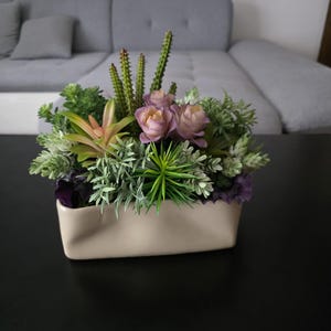 Puede incluir: Un arreglo floral artificial en una maceta rectangular beige. Contiene suculentas y flores verdes, moradas y rosas. El conjunto está colocado sobre una superficie negra, creando un efecto natural.