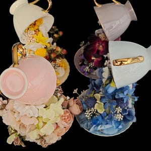 May include: Three upside-down teacups with floral arrangements inside. The cups are white, pink, and light blue with gold accents. The flowers are yellow, purple, and blue.