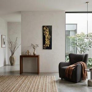May include: An interior shot of a modern living room with neutral tones. A dark gray armchair with a brown throw blanket sits near a window. A jute rug and a dark wood console table with decorative items are also visible. A black and gold abstract art piece hangs on the wall.