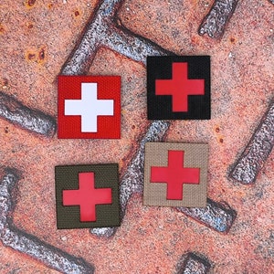 以下が含まれることがあります： 赤地に白十字、白地に赤十字、黒地に赤十字、ベージュ地に赤十字の刺繍パッチが4つ。パッチはさび付いた金属製の表面に置かれています。