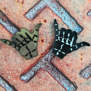 Könnte beinhalten: Zwei Stoff-Patches in Form einer Hand, die das Shaka-Zeichen zeigt. Ein Patch ist schwarz mit weißen Details und der andere ist grün mit schwarzen Details.