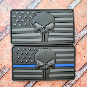 May include: Two black and gray patches featuring the American flag design with a skull emblem. One patch has a gray stripe, and the other has a blue stripe. The patches are rectangular and have a raised, textured design.