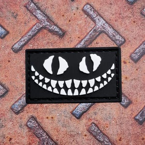 May include: Black rectangular patch featuring a white Cheshire Cat grin and eyes. The patch has a black border and is set against a textured, rusty metal surface. The design is a stylized, cartoonish representation of a cat's face.