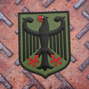 Peut inclure: Écusson noir et vert représentant un aigle stylisé avec des accents rouges sur le bec et les serres. L'écusson a une bordure noire et est posé sur un fond vert. Le fond est une surface métallique rouillée.