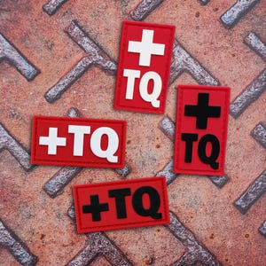 Op de afbeelding: Vier rechthoekige rode patches met de letters TQ en een kruissymbool. Twee patches hebben witte letters en een wit kruis, terwijl de andere twee patches zwarte letters en een zwart kruis hebben. De patches zijn voorzien van een gestikte rand.