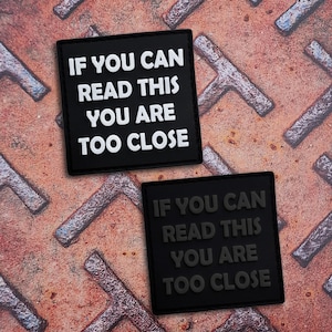 Puede incluir: Dos parches cuadrados con esquinas redondeadas. Uno es negro con texto blanco en relieve que dice IF YOU CAN READ THIS YOU ARE TOO CLOSE. El segundo es negro con texto negro en relieve que dice IF YOU CAN READ THIS YOU ARE TOO CLOSE.