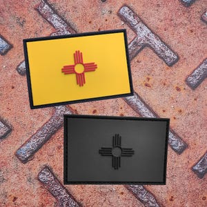 Könnte beinhalten: Zwei rechteckige Aufnäher mit der Flagge des US-Bundesstaates New Mexico. Der obere Aufnäher hat einen gelben Hintergrund mit einem roten Zia-Sonnensymbol, das schwarz umrandet ist. Der untere Aufnäher ist schwarz mit einem schwarzen Zia-Sonnensymbol, ebenfalls schwarz umrandet.
