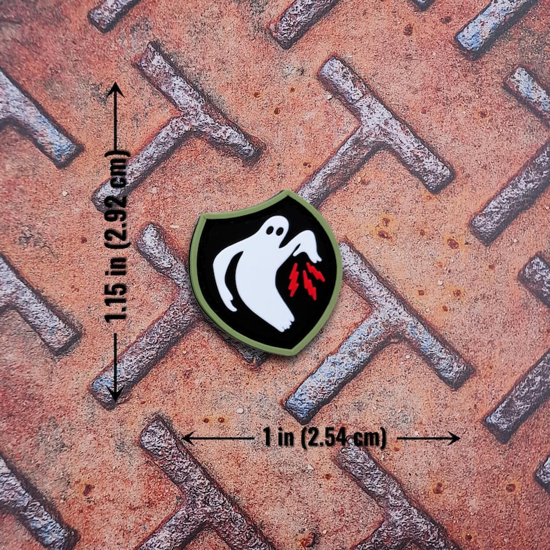 Op de afbeelding: Een schildvormige patch met een wit spookontwerp en rode vlammen op een zwarte achtergrond, omlijnd in groen. De patch is 2,54 cm breed en 2,92 cm hoog. Geplaatst op een roestig, getextureerd metalen oppervlak.