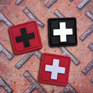 Könnte beinhalten: Drei quadratische Aufnäher mit Kreuzdesign. Einer ist rot mit einem schwarzen Kreuz, ein anderer ist schwarz mit einem weißen Kreuz, und der dritte ist rot mit einem weißen Kreuz. Sie liegen auf einer strukturierten, rostigen Metalloberfläche.