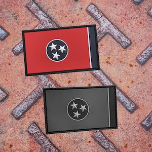 Op de afbeelding: Twee rechthoekige patches met het ontwerp van de vlag van de staat Tennessee. Eén patch heeft een rode achtergrond met een witte cirkel met drie witte sterren en een blauwe streep. De andere patch heeft een grijze achtergrond met hetzelfde ontwerp.