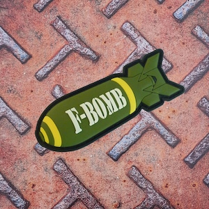 May include: A green and yellow patch in the shape of a bomb with the text "F-BOMB" in white. The patch is on a rusty, textured metal surface.
