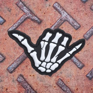 Puede incluir: Un parche en blanco y negro que representa una mano de esqueleto en el gesto shaka. El parche tiene un borde negro y huesos blancos. El fondo es una superficie metálica oxidada y texturizada.