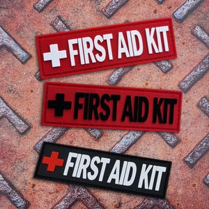 Könnte beinhalten: Drei rechteckige Aufnäher mit dem Text "FIRST AID KIT" und einem Kreuzsymbol. Der obere Aufnäher ist rot mit einem weißen Kreuz und weißem Text. Der mittlere Aufnäher ist rot mit einem schwarzen Kreuz und schwarzem Text. Der untere Aufnäher ist schwarz mit einem roten Kreuz und weißem Text.