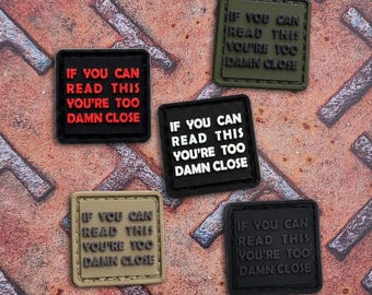 May include: Five square patches with the text IF YOU CAN READ THIS YOU'RE TOO DAMN CLOSE. One patch is black with red text, one is olive green with black text, one is black with white text, one is tan with black text, and one is black with black text.