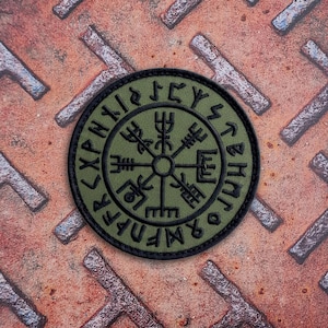 May include: A circular olive green patch featuring a black embroidered Vegvisir symbol at the center, surrounded by a ring of black runic characters. The patch has a thick black stitched border and a flat, fabric-like texture throughout.
