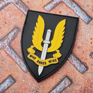 Könnte beinhalten: Schwarzer Aufnäher mit gelbem Flügeldesign und silbernem Schwert. Der Text "WHO DARES WINS" ist auf einem gelben Banner zu sehen. Der Aufnäher hat eine Schildform.