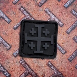 May include: A black square patch featuring a Jerusalem cross design. The patch has a raised, textured appearance and is set against a rusty, textured metal surface. The Jerusalem cross is a heraldic symbol.