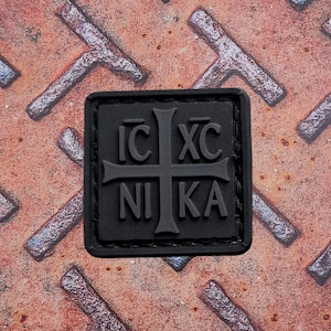 Pode incluir: Um patch quadrado preto com um desenho de cruz e as letras gregas IC XC NIKA. O patch &eacute; feito de um material emborrachado e est&aacute; sobre um fundo enferrujado e texturizado.