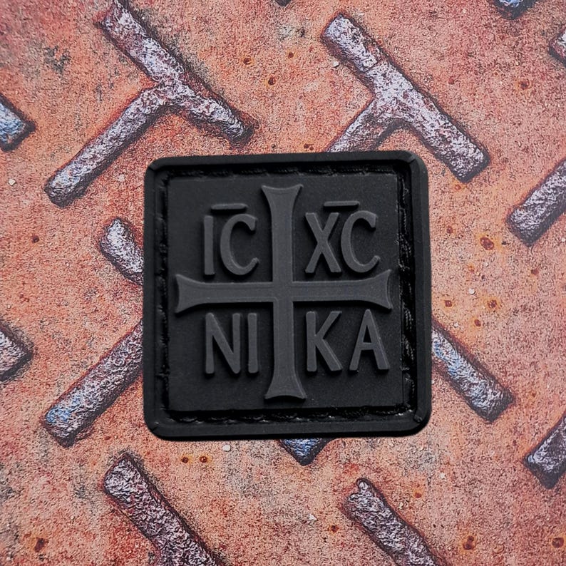 Pode incluir: Um patch quadrado preto com um desenho de cruz e as letras gregas IC XC NIKA. O patch &eacute; feito de um material emborrachado e est&aacute; sobre um fundo enferrujado e texturizado.