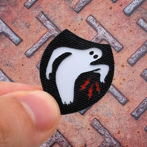 Könnte beinhalten: Ein schwarzer Aufnäher mit einem weißen Geisterdesign. Der Geist hat zwei schwarze Augen und rote Blitze, die aus seinem Mund kommen. Der Aufnäher wird in einer Hand gehalten.