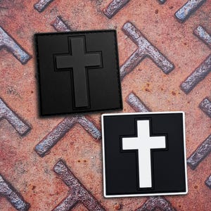 May include: Two square patches featuring a raised cross design. One patch is entirely matte black, while the other is black with a white cross. Both items have a clean, geometric border and a flat, uniform surface texture suitable for attachment.
