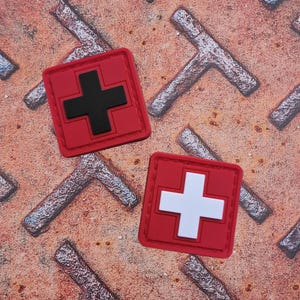 May include: Two square, red patches with a cross design. One patch has a black cross, and the other has a white cross. The patches are on a textured, rusty metal surface.