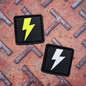May include: Two square black patches with a lightning bolt design. One patch has a yellow lightning bolt, and the other has a white lightning bolt. The patches are on a rusty, textured metal surface.